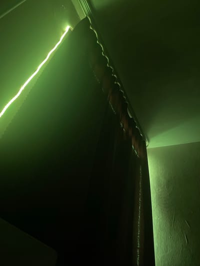 The image shows an interior view of a room with dark curtains partially drawn, illuminated by a bright green LED strip light. The light source runs along the top edge of the curtains, casting a vibrant green glow on the surrounding ceiling and walls. The curtains themselves appear to be a dark, possibly red or brown fabric, with a scalloped or ruffled edge visible near the top. The ceiling is a light color, reflecting the green light. A textured wall is visible to the right, also washed in the green illumination. The scene suggests an attempt to create ambient lighting, possibly in a residential or commercial setting. No people or specific actions are depicted. The location in Kyzyl, Russia cannot be determined from the visual information provided.