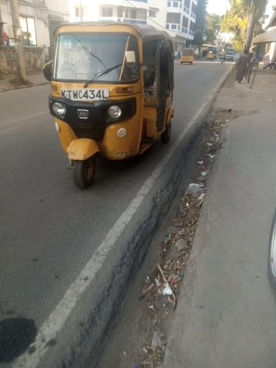 The image depicts a street scene within the Ganjoni/Shimanzi ward, Kenya, illuminated by natural daylight. A yellow auto-rickshaw (tuk-tuk) with Kenyan registration plate K TWC 434L is centrally positioned on an asphalt road, angled slightly towards the right, appearing either stationary or in slow motion. The vehicle features a black front grille with "RE" branding and shows minor scuff marks on its lower front bumper.
To the immediate right of the auto-rickshaw, a concrete gutter runs parallel to the roadway, filled with a substantial quantity of refuse including plastic bags, paper debris, and organic matter. Adjacent to the gutter is a paved sidewalk or shoulder. In the background, the road extends into the distance, populated by other vehicles including at least two more yellow auto-rickshaws, one motorcycle, and a light-colored van.
Multi-story buildings constructed of light-colored masonry line both sides of the street. On the far right side of the image, a person dressed in dark clothing is observed walking on a sidewalk, moving away from the camera. A portion of a wheel from another vehicle, likely the camera source, is visible in the extreme lower right foreground.
