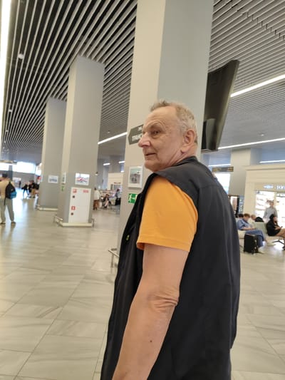 The image shows an older man with light-colored hair and fair skin, wearing an orange t-shirt and a dark vest. He is standing in what appears to be an indoor public space, possibly an airport terminal, given the tiled floor, modern ceiling design with integrated lighting, and pillars. In the background, other individuals can be seen, some sitting and some walking. There are also signs and what looks like information displays. The man is looking slightly to his right, with his body angled away from the camera. The overall lighting suggests an indoor environment. The location is identified as Khrabrovo, Russia, which is the location of Khrabrovo Airport (KGD) serving Kaliningrad.