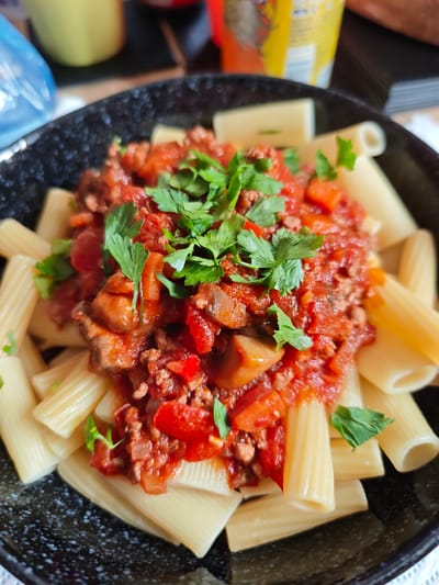 This media file is a close-up, overhead shot of a bowl of pasta. The pasta appears to be rigatoni, arranged in a circular pattern within a dark, speckled bowl. The pasta is topped with a rich, red meat sauce, likely a bolognese, which also contains diced tomatoes and mushrooms. Freshly chopped parsley is sprinkled generously over the sauce as a garnish.
The setting appears to be a casual indoor environment, possibly a dining table. In the blurred background, there are hints of other objects, including what looks like a yellow cup or container and a dark surface, perhaps a table. The lighting suggests it is daytime. No people are visible in the frame, and the focus is entirely on the food. The overall impression is one of a freshly prepared, hearty meal. There is no visible text in the image.