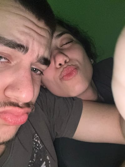 The image depicts two individuals in a close-up, chest-up shot. On the left, a male subject with light-colored eyes, prominent eyebrows, and a light mustache looks directly at the camera with pouted lips. He wears a dark gray t-shirt with a light graphic. On the right, a female subject is positioned closely beside him, her head resting near his shoulder. She has dark hair, closed or squinting eyes, and pouted lips, mirroring the male's expression. A small, shiny earring is visible in her right ear. She wears a dark-colored shirt. The background is a uniform dark green. No visual information within the image indicates the city of Arkovna, Bulgaria.