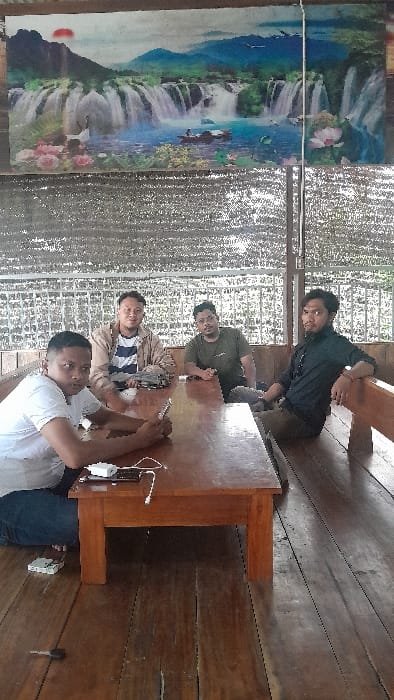 Four adult males are depicted seated around a dark brown rectangular wooden table within an open-sided structure, likely a public eatery, located in the city of Aluebilirayeuk, Indonesia. Above, a mesh tarp serves as a canopy, and a large banner displaying a vibrant landscape with waterfalls, mountains, water, flora, a crane, a boat, and a red sun is affixed to the wall behind them. The floor is constructed from wooden planks.
From left to right:
1. A male in a white t-shirt and blue jeans kneels beside the table, holding a black smartphone and looking down at it. A white power bank connected to a charging cable rests on the table in front of him. A white smartphone or device is on the wooden floor near his left foot.
2. A male wearing a light brown jacket over a striped t-shirt sits on a wooden bench behind the table, resting his right arm on the table's surface and looking towards the camera.
3. A male with glasses, wearing an olive green t-shirt, sits on the wooden bench, with his forearms on the table, looking towards the camera.
4. A male with dark hair and a beard, dressed in a black long-sleeved shirt and dark pants, sits on the wooden bench, leaning back slightly with his right arm on the bench's backrest, and looks towards the camera.
A silver key is visible on the wooden floor near the lower left table leg. Green foliage is visible beyond the structure's open side.
