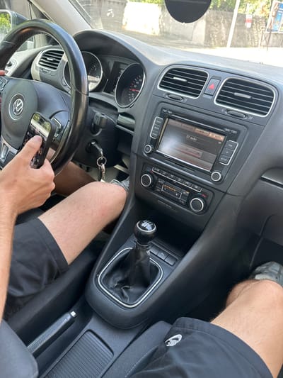 An interior shot from the passenger side of a car, looking towards the driver's seat and dashboard, captures a person's hand holding a smartphone. The car appears to be a Volkswagen, indicated by the logo on the steering wheel. The dashboard features a radio display showing "BT audio," climate control knobs, and air vents. A gear stick is visible in the foreground. The scene suggests the car is parked or stationary, given the driver's focus on the phone and the presence of a key in the ignition. The exterior environment, glimpsed through the windshield, shows a bright, outdoor setting with some vegetation and buildings, hinting at a location in Bagard, France. The image is a close-up, focusing on the car's interior and the interaction with the phone.