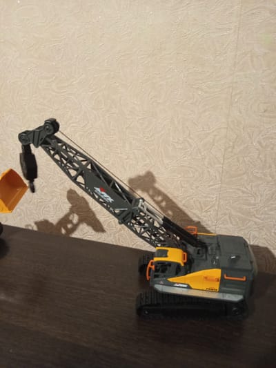 An image shows a toy construction crane with a yellow bucket, set against a textured wallpaper. The crane is gray and yellow, with black treads and a movable boom. A shadow of the crane is cast on the wallpaper. The toy is placed on a dark wooden surface. There are no people or animals in the image. The scene is indoors and appears to be in a residential setting in Novotroitsk, Russia.