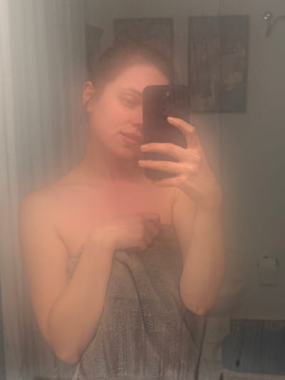 A woman is taking a selfie in a bathroom mirror, holding a black smartphone with a triple-lens camera in her right hand. She appears to be wrapped in a gray towel. The bathroom features a toilet paper dispenser, artwork on the wall, and a bathroom sink. It is not possible to determine if the location is in Perugia, Italy, from the image alone.