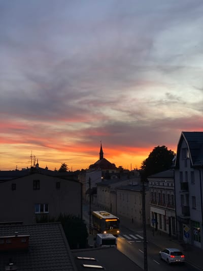 This is an eye-level, high-angle shot of a street scene in Swarzędz, Poland, at sunset. The sky is filled with vibrant hues of orange, pink, and purple, with streaks of lighter gray clouds. In the background, the spire of a church is visible, silhouetted against the colorful sky. The street is lined with multi-story buildings, some of which appear to be residential, while others have storefronts. A yellow bus, with its headlights on, is driving down the street, and a white van is parked on the side. A white car is also visible driving away from the camera. The street has a pedestrian crossing. The image captures a peaceful evening in a Polish town.