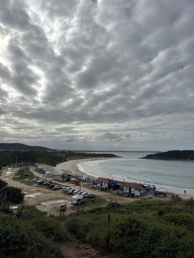 The image captures a wide aerial view of a curved sandy beach along a coastline, identified as Cabo Frio, Brazil. The dominant feature of the sky is a sprawling expanse of gray and white clouds, forming distinct wave-like patterns across the frame.

The scene depicts a long stretch of light-colored sand meeting tranquil, light blue-green ocean waters with gentle waves breaking near the shore. In the distance, the coast extends, eventually meeting a headland or island.

Along the immediate coastline, set back from the water, there's a bustling area of beach structures. These include numerous small, low-rise buildings, likely beach shacks, kiosks, or restaurants, many featuring awnings (some blue) and white tents. Tables and chairs are visible outside some of these establishments. Notable visible text includes "RESTAURANTE" and "PEIXARIA" on different buildings, indicating dining and fish sales.

Adjacent to these structures, a dirt path or parking area runs parallel to the beach, filled with a line of parked cars, predominantly white, and possibly some motorcycles or quad bikes. On the right side of the image, a steep, verdant slope or cliff covered in dense green vegetation and trees frames the beach area. Scattered small figures are visible on the beach and in the water, suggesting people are present, likely enjoying the beach despite the overcast conditions.

The time of day appears to be daytime, under cloudy skies that diffuse the light, preventing harsh shadows. The overall impression is that of a popular beachside settlement.