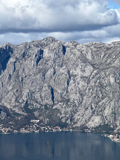 The image presents a panoramic view of a coastal region near Bogdašići, Montenegro, captured from an elevated perspective. A formidable, craggy grey limestone mountain range, exhibiting sparse dark green or brown vegetation, forms the primary backdrop, rising steeply from the water. The sky above is largely overcast with dense grey clouds, though a segment of clear blue sky is visible in the upper right. In the middle ground, a dark blue body of water, appearing as a bay or inlet, mirrors the subdued light. Along the narrow coastal strip at the base of the mountains, a town or village is visible, characterized by numerous white-walled buildings with red or terracotta roofs, densely clustered along the shoreline and extending slightly up the lower mountain slopes. Within the bay, multiple geometric patterns of structures consistent with marine aquaculture are discernible. In the immediate foreground, two slender, out-of-focus, bare plant shoots protrude from the bottom of the frame. No individuals, actions, or direct interactions are visible.