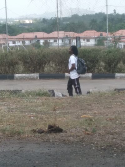 The scene depicts an outdoor environment, located in Osu, Nigeria, under an overcast sky. In the midground, a lone individual, appearing male with long dark dreadlocks, walks rightward across a paved surface, looking to the right. The individual wears a white t-shirt, dark trousers with a white stripe on the left leg, and carries a dark backpack. Immediately behind the person is a concrete curb painted black and white. Behind the curb, a dense line of green bushes spans the width of the image. The foreground consists of dry, patchy dirt and sparse grass, with a dark, irregular pile of organic matter present on the ground. In the background, a wire fence, likely chain-link, is visible. Behind this fence are several one-story buildings featuring reddish-brown tiled roofs. Further in the distance, dense green foliage, including palm trees, and hazy hills are discernible. A tall, slender metal tower is partially visible on the right side of the background. No other individuals or active interactions are observed.