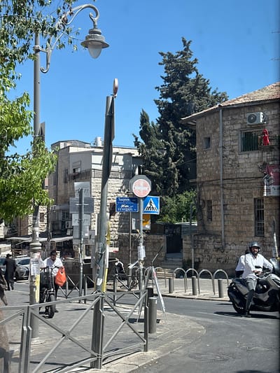 This is a vertical, eye-level shot of a street scene in al-Eizariya, Palestinian Territories. The image depicts a bright, sunny day with a clear blue sky. In the foreground, a light fixture with ornate detailing hangs from a pole, partially obscured by lush green trees. A street sign with Hebrew and English text is visible, reading "YEHEZKEL ST." Further down the street, a person on a motorcycle, wearing a white shirt and a helmet, is seen. The buildings in the background are made of stone, characteristic of the region. The overall scene conveys a sense of daily life in a Middle Eastern urban environment. The time of day appears to be midday, given the strong sunlight and shadows.