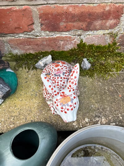 The image depicts an outdoor scene in an unspecified location. A patterned ceramic rabbit ornament sits on a rough concrete surface adjacent to a brick wall covered with moss. Nearby, a section of a metal bucket containing water and a green watering can are visible. There are no people present and no actions or interactions are occurring. The environment suggests a garden or outdoor setting. There is no indication it is in Shrewsbury, United Kingdom.