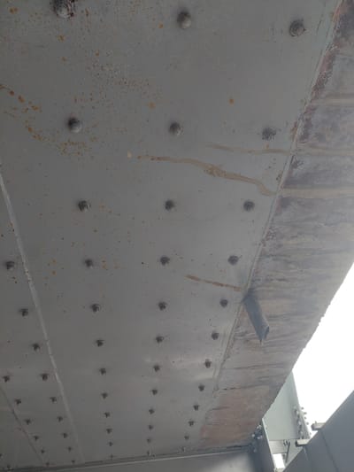 The image shows the underside of a gray, riveted metal structure, possibly a bridge or overpass, in Parit Baru, Indonesia. The metal is weathered and shows signs of rust and water stains. Numerous rivets are visible, arranged in a grid pattern. A dark, cylindrical pipe protrudes from the edge of the structure on the right side of the frame. The sky, visible in the upper right corner, appears overcast and bright. The perspective is looking upwards at the underside of the structure.