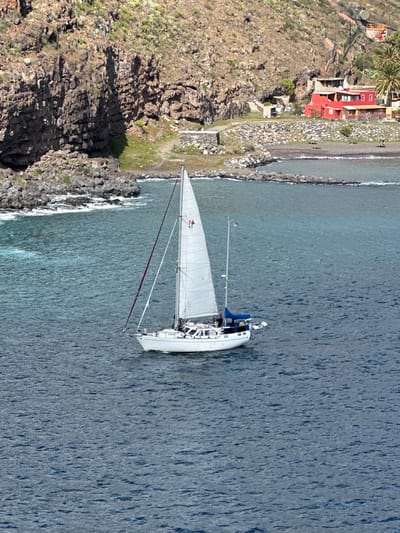 A white sailboat with its main sail unfurled navigates calm, blue-green waters in the vicinity of San Sebastián de la Gomera, Spain. The sail bears a red insignia, possibly "M 43." A German flag flies from the stern, where a person in dark attire is visible in the cockpit area under a blue dodger. The boat features a white hull and visible rigging. The background consists of a rugged, rocky coastline composed of dark brown and reddish-brown cliffs on the left, exhibiting sparse green vegetation. Towards the center-right, a small, dark pebble beach is visible, protected by two stone breakwaters extending into the water. A dirt path leads from the beach area uphill. Nestled on the hillside above the beach are several structures, most notably two buildings with distinctive red-orange exteriors and brown roofs, surrounded by some green foliage, including palm trees. The scene is illuminated by bright daylight, indicating clear weather.