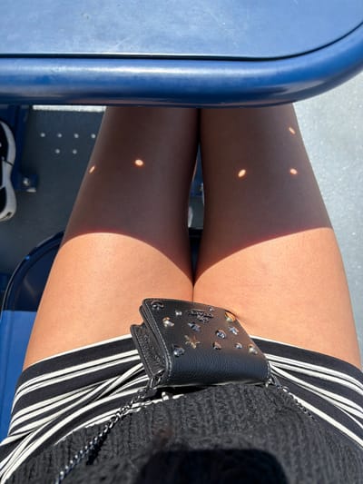 The image shows a close-up view of a person's legs and a wallet. The person is sitting on a blue seat, as evidenced by the blue structure at the top of the image and the blue seating visible on the left. Sunlight is filtering through, creating dappled shadows on the person's legs. The person is wearing a black and white striped skirt or shorts, and a black knitted top. A small black wallet, adorned with silver studs and stars, is placed on the person's lap. The background appears to be a textured surface, possibly a wall or ground, with small, bright circular reflections. The scene is shot from a high angle, looking down at the person's legs. There is no identifiable information about the location being in Southern Gulf Islands Electoral Area, Canada within the image itself.
