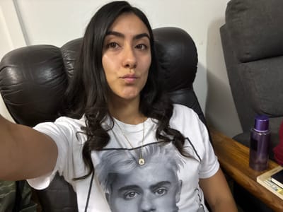 A young woman with dark, wavy hair is seated in a dark leather armchair, facing the camera. She is wearing a white t-shirt with a black and white image of a person's face printed on it, and a gold necklace with a circular pendant. She has her lips pursed. To her right, on a wooden table, is a tall purple bottle and some books. The background consists of a white wall and a grey upholstered chair. The image appears to have been taken indoors, possibly in a home or living room setting, and there is no indication of its location in Imatra, Finland.