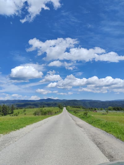 The image presents a straight asphalt road stretching into the distance under a vibrant blue sky dotted with cumulus clouds. The road appears to lead toward a range of forested mountains. The landscape on either side of the road consists of green fields, interspersed with patches of trees and shrubs. A fence can be seen enclosing one of the fields on the right side of the road. The scene is illuminated by bright daylight. A small, reddish object, possibly a bird or drone, is visible in the upper left quadrant of the sky. This is a rural scene, indicative of a location such as Kasak, Bulgaria.