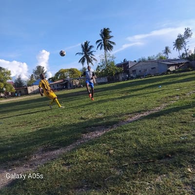 The image captures two men playing soccer on a grassy field in Nungwi, Tanzania, located at coordinates -5.726 latitude and 39.301 longitude. The field is surrounded by trees and buildings, with a few spectators watching the game from the sidelines. The men are wearing soccer jerseys, and the one in the foreground is kicking a ball in the air. There are a few palm trees in the background, providing a natural backdrop for the game. The sky is clear and blue, indicating a bright, sunny day. The overall scene appears to be a casual soccer game taking place in a relaxed, community setting.