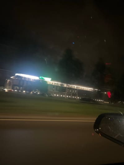 The image shows a long, multi-story building illuminated by lights at night, with a green neon sign on its roof. The building appears to be a hotel or a commercial establishment. The scene is captured from a moving vehicle, as evidenced by the blurriness and the presence of a car's side-view mirror in the foreground. There is a road in front of the building, and grassy areas with trees behind it. The sky is dark, indicating nighttime. The overall impression is a view of a lit-up building from the perspective of someone driving past. The location is Krivina, Bulgaria.