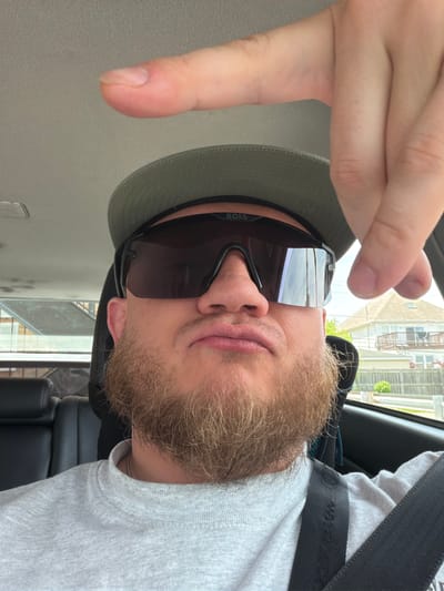 The image depicts a person in a vehicle, likely a car, as suggested by the interior view. The individual is wearing sunglasses with "BOSS" branding, a light-colored hat, and a gray shirt. A seatbelt is visible across their chest. The person's beard is light brown. Their hand is raised, with fingers spread.
Outside the vehicle, blurry shapes suggest a cityscape with buildings, possibly apartments or office structures, with some greenery visible. While difficult to pinpoint the exact location within Morton Grove without more distinct landmarks, the general architectural style in the background suggests an urban or suburban environment consistent with parts of Morton Grove, Illinois.