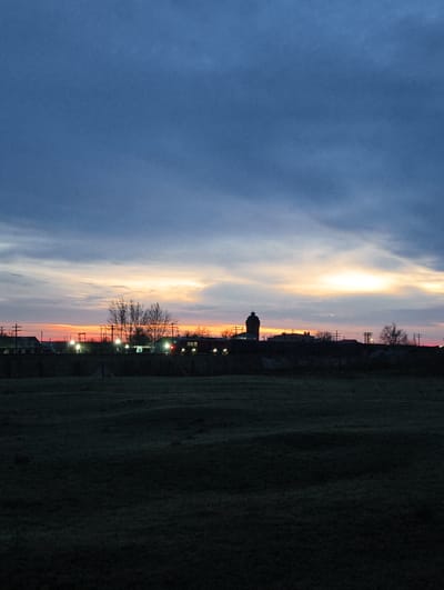 The image presents a twilight scene in the Достық ауылдық округі, Kazakhstan. The foreground is dominated by a dark, grassy field. Beyond the field, a cluster of buildings and structures are silhouetted against a colorful horizon, with shades of orange and yellow giving way to a blue and cloudy sky. There are utility poles and bare trees interspersed among the buildings, and a prominent, dark, tower-like structure stands out from the skyline. Lights are visible within the buildings, suggesting activity despite the fading light.