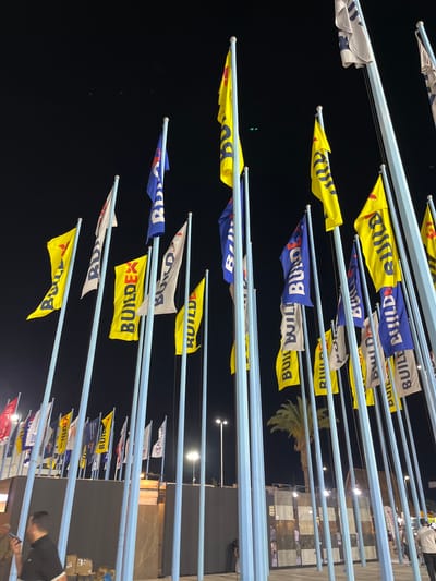 The image shows an outdoor event at night in Al-Ghizlaniya, Syria. A multitude of flags are displayed on tall poles. Many of the flags are yellow and blue, with text that appears to be "BUILDEX" and "BUILD". Some white flags with blue lettering are also present. In the background, there are buildings and lighting fixtures. Several people are visible in the foreground, some walking and one standing while looking at their phone. The environment appears to be a public space or exhibition area, with a palm tree visible in the mid-ground.