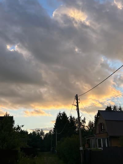The image depicts a rural landscape, likely within the city of Слава Снт, Russia, under a dramatic sky filled with a mix of dark and bright, sunlit clouds. In the foreground, to the right, a wooden house with a dark-pitched roof is visible. The house has a porch with large windows. A dark fence runs along the front of the property. To the left of the house, dense green trees form a line. A dirt path leads away from the viewer into the distance, bordered by vegetation and trees on either side. A utility pole with wires stretches across the upper portion of the image, with a second, thinner wire also visible. The overall impression is of a cloudy sunset in a semi-rural or dacha setting.