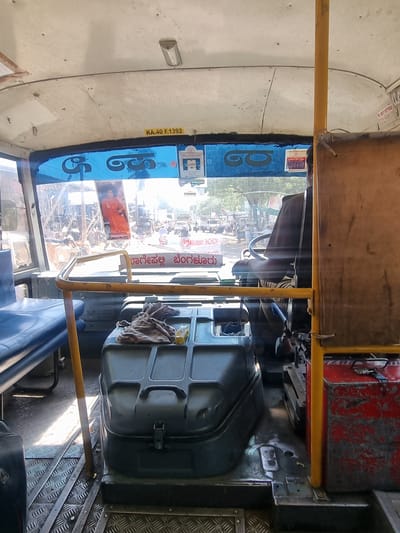 This image captures the interior of a public bus in Gorantla, India, viewed from a forward-facing perspective, looking through the windshield onto a street scene.

The bus interior features a light-colored, possibly off-white, ceiling with visible rivets and a single rectangular overhead light fixture. A yellow sticker with "KA.40 F.1392" is affixed to the ceiling above the windshield. A blue-tinted band runs across the top of the windshield, displaying prominent Kannada script, including "ಹಾಗಲೂರು ಬೆಂಗಳೂರು" (Hagaluru Bengaluru), indicating a bus route. Various smaller stickers, some with additional Kannada text and dates, are also present on the windshield and the blue band. A large image of a person in an orange robe, likely a spiritual leader, is prominently displayed in the center of the windshield.

To the left, a blue-cushioned bench seat is visible. In the center, a large, dark grey or black engine cover/console dominates the floor space between the passenger area and the driver's seat. A crumpled brown cloth and a small yellow bottle rest on top of this console. The floor on the left has a silver-colored, diamond-plate texture, transitioning to a darker, worn surface towards the driver's side. A vertical yellow support pole stands on the right, near a red, weathered metal box with visible wiring and a sticker reading "FC KIT FITTED 11-12-23". A horizontal yellow bar extends from the left toward the central console.

A person, presumably the driver, is partially visible from behind on the right, wearing a dark garment, with hands on the steering wheel.

Through the windshield, a busy street scene unfolds under bright daylight. Numerous pedestrians are visible, along with multiple vehicles including cars, motorcycles, and what appear to be auto-rickshaws, suggesting an active urban or semi-urban environment. Green foliage from trees is discernible in the background. The scene indicates the bus is either stopped in traffic or at a bus stop along an active route.