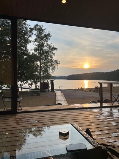 This image captures a serene sunset view of a lake from a covered wooden deck, likely in the vicinity of Hämeenlinna, Finland. The foreground features a wet wooden deck with reflections of trees and the sky. A glass-topped table with a small box and a smartphone rests on the deck. Beyond the deck, a sandy area leads to a lake with a wooden pier. Several people, including children, are visible in and near the water, suggesting a recreational area. The setting sun casts a warm golden glow across the water and the sky, illuminating a treeline in the distance. The overall atmosphere is peaceful and tranquil.