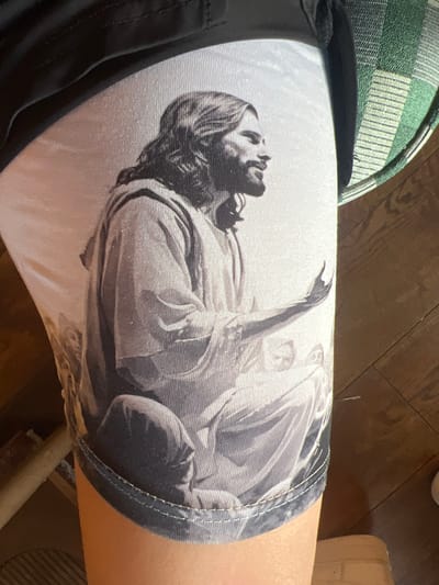 The image shows a person wearing a pair of shorts with an image of what appears to be Jesus Christ raising his arm printed on them. The shorts are white and the image is in black and white. Part of the person's leg is visible, as well as a chair with a green and white patterned cushion. The floor beneath the chair is wooden. The location is not discernible from the image. The image does not contain any information about Bristol, United Kingdom.