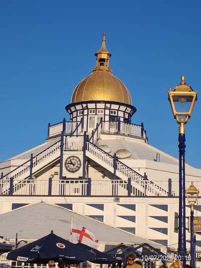 The image captures a close-up view of the Eastbourne Pier in Eastbourne, United Kingdom, under a clear blue sky. The iconic golden dome of the pier's pavilion is prominently featured at the top, adorned with a small red finial. Below the dome, a white circular structure with blue trim and railing surrounds the pavilion, featuring external staircases. A large clock with Roman numerals is visible on one of the lower sections of the pavilion. A tall, ornate golden lamppost with a blue base stands to the right of the pavilion, partially obscuring the dome. In the foreground, a blue umbrella with a "Camden Town Brewery" logo is partially visible, along with an English flag. The bottom right corner of the image displays a date and time stamp: "10/07/2026 20:13". Several people are visible in the lower part of the frame, suggesting a public setting, possibly an entertainment venue or restaurant area on the pier. The architecture is characterized by white painted structures with blue accents and decorative elements.