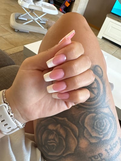 The image shows a close-up of a person's hand with freshly done French tip acrylic nails. The nails are long and square-shaped, with a pink base and white tips. The person's arm, with visible tattoos of roses, is also in the frame. A white smartwatch with a silver chain bracelet is on the person's wrist. In the background, a white baby walker and a glimpse of what appears to be a TV screen and cabinet can be seen. The floor tiles suggest an indoor setting. The lighting indicates it's daytime. No specific emotions are conveyed, and no text is legible in the image. The location is identified as Veghel, Netherlands.