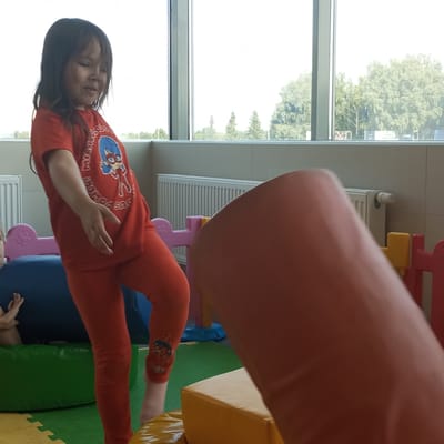 A young girl wearing a red t-shirt with a Miraculous Ladybug graphic and red leggings is actively engaged in play in an indoor children's recreation area. Her left leg is bent and raised, suggesting she is climbing or maneuvering over an obstacle, which appears to be a large, orange cylindrical object positioned in the foreground. The girl's expression is one of concentration or slight strain.
Behind the girl, various play structures are visible, including a blue padded mat and a pink decorative element with flower-like cutouts. To the right, a yellow foam block and part of a red play fence can be seen. In the background, a white radiator is mounted on a tiled wall. Large windows dominate the upper portion of the image, offering a view of trees and buildings, suggesting an outdoor environment beyond the indoor play space. The overall setting is brightly lit, likely due to the windows.
The image was captured in Naberezhnye Chelny, Russia.