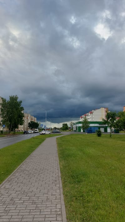The image captures a scene in the city of Masty, Belarus. A paved pedestrian walkway runs along the right side of a road, bordered by a neatly mown grassy area. On the left, a multi-story apartment building lines the road, with a tall green tree in front of it. Further down the road, several cars are parked. On the right side of the road, across from the apartment building, is a low-rise building with a green facade, possibly a store or commercial establishment. Beyond this building, more apartment buildings are visible. The sky is filled with dark, heavy storm clouds, creating a dramatic and overcast atmosphere.