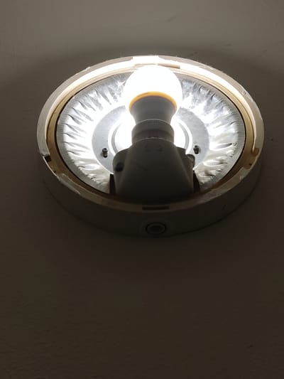 A close-up, high-angle shot captures a ceiling light fixture with a lit LED bulb. The fixture is circular with a white outer casing and a ribbed, reflective interior surface. The LED bulb, white and cylindrical with a brown base, is mounted in a white socket. The ceiling around the fixture is painted a neutral color, possibly beige or off-white, and appears slightly textured. The lighting creates a soft glow and casts subtle shadows. No people or other objects are visible in the frame. The image is focused on the light fixture.