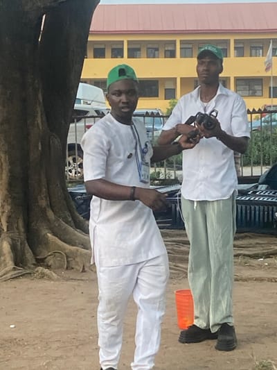 Two men, likely Nigerians, wear green hats and white clothing. The man in the foreground stands near a tree and wears a white outfit, while the man behind him wears a white shirt and light green trousers. Both men appear to be photographers, each holding cameras. The background features a building with multiple windows and a light yellow color. A car is also visible behind the men.