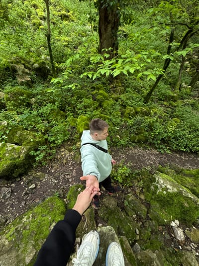 A high-angle shot captures a man standing on a dirt path in a green forest. The man is wearing a light green sweatshirt and black pants with black shoes. He is holding hands with someone whose arm and part of their legs and white sneakers are visible on the right. The environment is lush with green moss covering rocks and trees. The dirt path meanders through the scene. This scene occurs in Tsabal, Abkhazia.