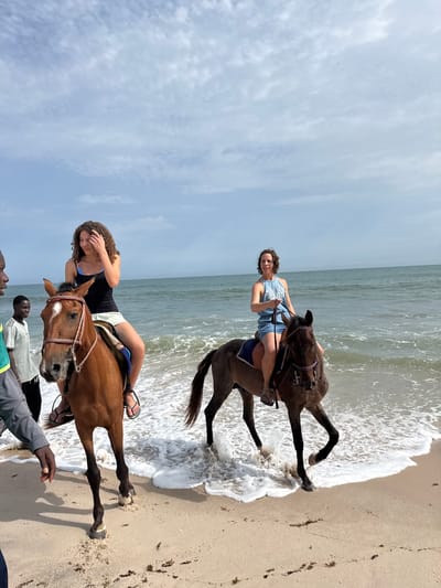 Two women are riding horses on a sandy beach in Mopo-Akinlade, Nigeria. The water line of the ocean is reaching the horses' legs. The sky is overcast. Several people are standing on the beach near the horse in the foreground.