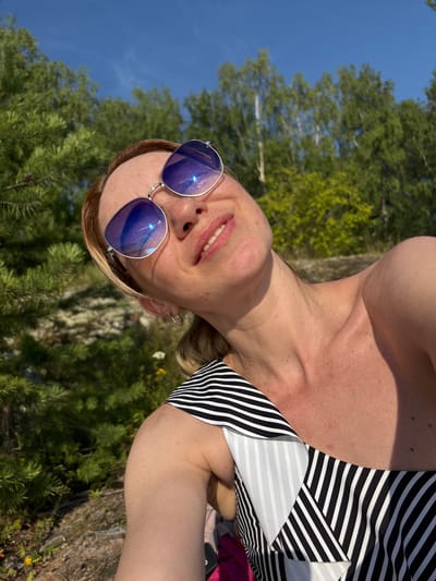 The image shows a woman wearing sunglasses and a striped swimsuit, outdoors with trees and a blue sky in the background. She appears to be taking a selfie, with her head tilted back and her mouth open in a smile. The environment suggests a natural setting, possibly a park or forest. There is no indication of the city of Shuiskaya Chupa, Russia, in the image or its context.
