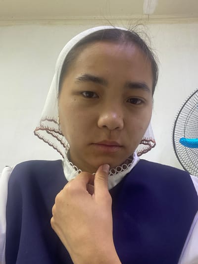 A young woman, wearing a white head covering with a decorative lace trim and a dark blue uniform with a white collar, is pictured in a medium close-up. Her right hand is raised, with her thumb and index finger lightly touching her chin, and her other fingers are tucked under her jawline. She is looking directly at the camera with a neutral expression. The background is a plain, lightly textured wall, and to the right of the woman, a portion of a black and silver electric fan with a bright blue element is visible. The lighting appears to be overhead, casting soft shadows. The scene is indoors, suggesting a domestic or institutional setting. There is no information to indicate this photo was taken in Kyzyl-Kyya, Kyrgyzstan.