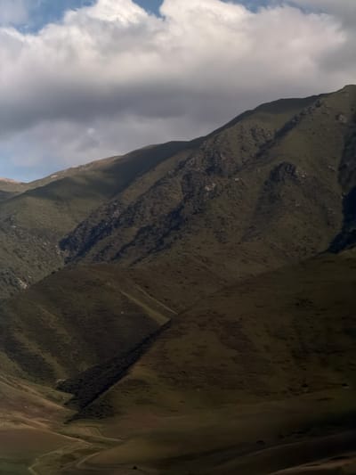The image depicts a dramatic, tilted view of a mountainous landscape, likely Karagai Bulak, Kyrgyzstan. The dominant subject is the rugged terrain, characterized by steep, dark slopes covered in vegetation. These slopes are crisscrossed by deep shadows, suggesting a time of day when the sun is casting long shadows, perhaps late afternoon or early morning. The texture of the ground appears rough and uneven, with scattered rocks and patches of earth.
A striking feature of the scene is the contrast between the dark mountainside and the bright, cloudy sky. Large, white clouds dominate a significant portion of the frame, with glimpses of blue sky peeking through. This suggests dynamic weather conditions, possibly a partly cloudy day. The clouds are illuminated from above, further emphasizing the time of day and the presence of sunlight.
There are no discernible people or man-made objects in the frame. The focus is entirely on the natural, imposing grandeur of the mountains. The overall mood conveyed is one of wild, untamed nature. The extreme tilt of the camera adds a sense of disorientation or a unique perspective, as if the viewer is looking up at the mountain from a precarious position or experiencing a moment of dramatic ascent or descent. No visible text is present in the image.