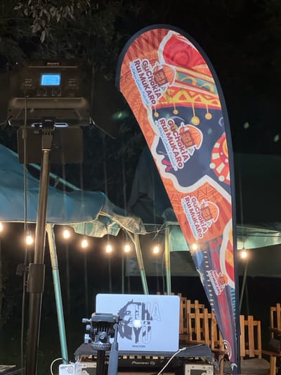 This media file captures an outdoor evening or nighttime event in Mururiini, Kenya, centered around a cultural podcast. The scene is illuminated by string lights and a powerful LED light panel, indicating it's dark outside.
The most prominent subject is a large, crescent-shaped banner adorned with vibrant, traditional-looking patterns in shades of red, orange, yellow, and blue. Repeated across the banner is the text: "GuChokiA Rui MuKARO - A Kikuyu Culture Podcast." The logo accompanying the text appears to be a stylized representation of a traditional hut or cultural headwear.
Below the banner, various equipment is visible. An open laptop, displaying a distinct black and white graphic with the stylized text "AHA" and "WACIURI," rests on a tripod. Adjacent to the laptop, the words "Pioneer DJ" are discernible, suggesting audio mixing or performance capabilities. A professional LED light panel, showing "100%" brightness on its digital display, is also mounted on a stand, providing bright illumination to the setup. In the background, stacked wooden pallets create a rustic backdrop, and draped fabric or tarp material suggests a temporary outdoor structure or stage. Dark foliage is visible in the furthest background, confirming the outdoor setting.
The arrangement of the banner, lighting, and audio equipment strongly implies a live recording session, a cultural performance, or a gathering associated with the "Kikuyu Culture Podcast." The artificial lighting confirms the event is taking place after sunset.