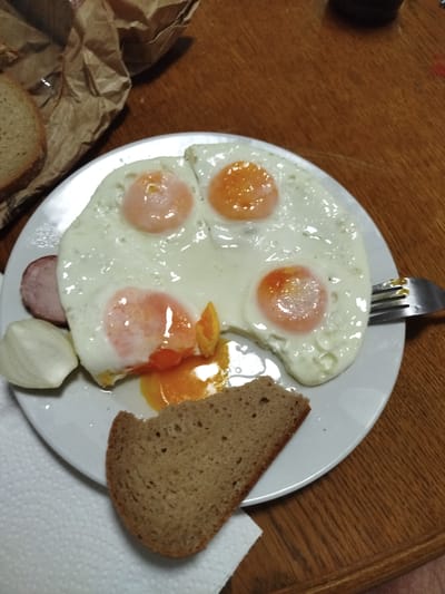 This image shows a plate of fried eggs with a slice of bread, a piece of ham, and a piece of onion on a wooden table. The eggs are cooked sunny-side up, with the yolks still runny. The bread is dark rye bread. The ham is a round slice of cured meat. The onion is a white slice. The meal is set on a white plate, with a white napkin underneath. A crumpled brown paper bag is in the background. A fork is visible on the right side of the plate, partially submerged in the egg whites. The lighting suggests this is an indoor setting, likely a home. There is no visible text in the image. The overall impression is of a simple, hearty breakfast or light meal.
