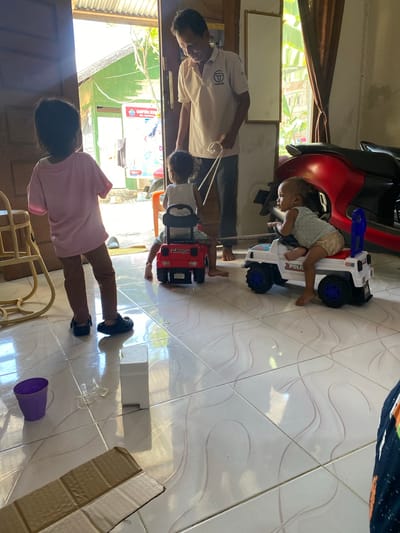 An adult male, wearing a light-colored polo shirt with a circular logo, stands in a doorway, looking down. To his left, a young child in a pink shirt and brown pants stands facing away from the camera, looking out the doorway. In front of the child, a red toy Jeep is visible, with another child sitting in it. To the right, a baby sits in a white toy police car. A red motorcycle is partially visible behind the toy cars. The scene appears to be indoors, with tiled flooring and sunlight streaming in from the doorway. The background suggests a building with a corrugated metal roof and a green wall. The image is set in Tuha Lada, Indonesia.