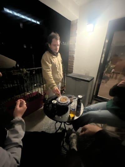 The image depicts an outdoor night scene on a balcony or patio in Amiens, France, featuring three individuals gathered around a small table.
In the center, a male subject with brown hair, wearing a light tan jacket and white trousers, stands facing the table. He is leaning slightly forward, with his right hand extended towards a blue and white patterned bowl filled with light-colored snacks or nuts.
The black, round, metal table with a textured surface is positioned in the foreground. On the table, in addition to the bowl, are two dark beverage cans, a clear plastic bottle containing a yellowish liquid, and a dark-colored smartphone. A small, dark-colored object, possibly an ashtray, is also present.
To the immediate left of the table, a person's left arm and hand are visible, holding what appears to be a cigarette or similar slender object. This individual wears a light grey sleeve. To the right of the table, a seated person's lower body is visible, wearing light blue ripped jeans and dark footwear.
The background features a dark night sky with distant building lights horizontally across the top. To the right, an exterior wall segment includes a section of red brick, a plain light-colored wall, and a bright, square outdoor light fixture illuminating the immediate area. A dark-framed glass door or window reflects ambient light. Below the light, a grey plastic outdoor storage cabinet is visible. A dark metal railing with a long red planter box containing some greenery is present on the left side, separating the balcony from the exterior. A patterned grey and white rug or mat covers the floor beneath the table.