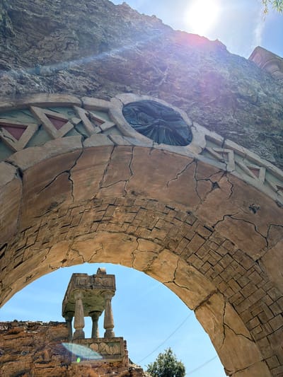 The image depicts an outdoor scene under a bright sky, likely in an amusement park or resort setting, given the artificial, weathered appearance of the structures. The primary focus is a large, cracked stone archway, viewed from below and looking upwards. The arch is constructed from textured, brown, stonelike material with visible faux cracks and an inset rectangular pattern along its upper edge, featuring triangular decorative elements. Within the arch, the bright sunlight flares.

Beyond the arch, through its opening, a clear blue sky is visible. In the distance, a small, ornate stone structure resembling a ruined altar or monument stands on a rocky ledge. This structure is supported by four fluted columns and features decorative carvings. The rocky ledge itself appears to be a man-made facade, with a rough, textured surface. A tree with green foliage is visible in the lower right portion of the frame, partially obscured by the arch. The overall impression is one of ancient ruins or a historically themed environment. This specific archway is part of the entrance to the Gardaland Magic Halloween area in Castelnuovo del Garda, Italy.