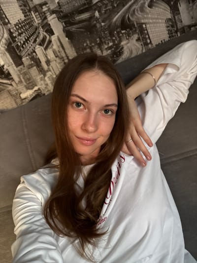 The image is a close-up selfie of a young woman with long brown hair, wearing a white long-sleeved shirt. She is lying on a grey sofa, with her left arm propped up behind her head. A mural of a city skyline is visible on the wall behind her. The lighting suggests an indoor setting, likely a home. There is no indication of the specific location within Novaya Ivanovka, Russia.