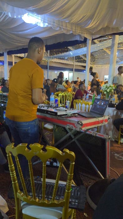 An indoor event with people at tables. A DJ in a yellow shirt and jeans stands behind a Pioneer DJ setup. There's a laptop on the DJ table, and a sound mixer is placed on a gold chair. The event space is decorated with white and blue draping.
