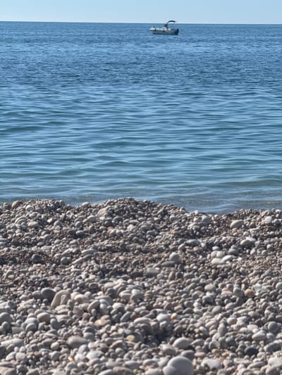 The image shows a beach scene in Vinaròs, Spain. A pebble beach is on the right, and the sea is on the left. Small waves are visible in the water. A boat is docked in the distance. The sky is clear and blue.