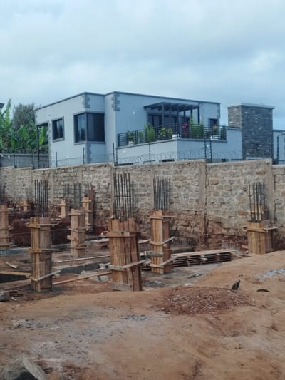 The image shows a construction site in Othaya, Kenya, located at latitude -0.44 and longitude 36.958. The site features a new residential building with a stone wall constructed around it. The building is two stories high, with a roof terrace. The exterior of the building is light gray, with large windows. There are several wooden posts in the foreground of the image, suggesting the building is under construction. The ground is a mixture of dirt, rocks, and concrete. There is a bird on the ground in the lower right of the image. The site is secured with a metal fence.