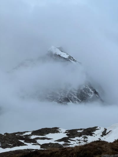 The image captures a dramatic, high-altitude mountain landscape, likely in the Alps near Poschiavo, Switzerland. The scene is enveloped in thick clouds or fog, creating an ethereal and monochromatic atmosphere.

In the foreground, on the right side of the image, a steep, rugged rock face is prominently featured. This cliff face is covered in irregular patches of white snow and dark, dry, coarse vegetation, indicating a harsh, cold environment. The perspective suggests the viewer is looking down or across from a high vantage point.

Through the shifting clouds in the middle ground and background, two distinct, jagged mountain peaks emerge. These peaks are also snow-dusted and rocky, appearing formidable and partially obscured by the atmospheric conditions. The fog creates a sense of depth and mystery, revealing only glimpses of the distant terrain.

The overall color palette is dominated by cool grays, whites, and dark browns, reflecting the overcast weather and snow-covered terrain. There is no visible sun, suggesting an overcast day. No human activity, structures, or visible text are present in the image. The scene conveys a sense of wildness, isolation, and natural grandeur.